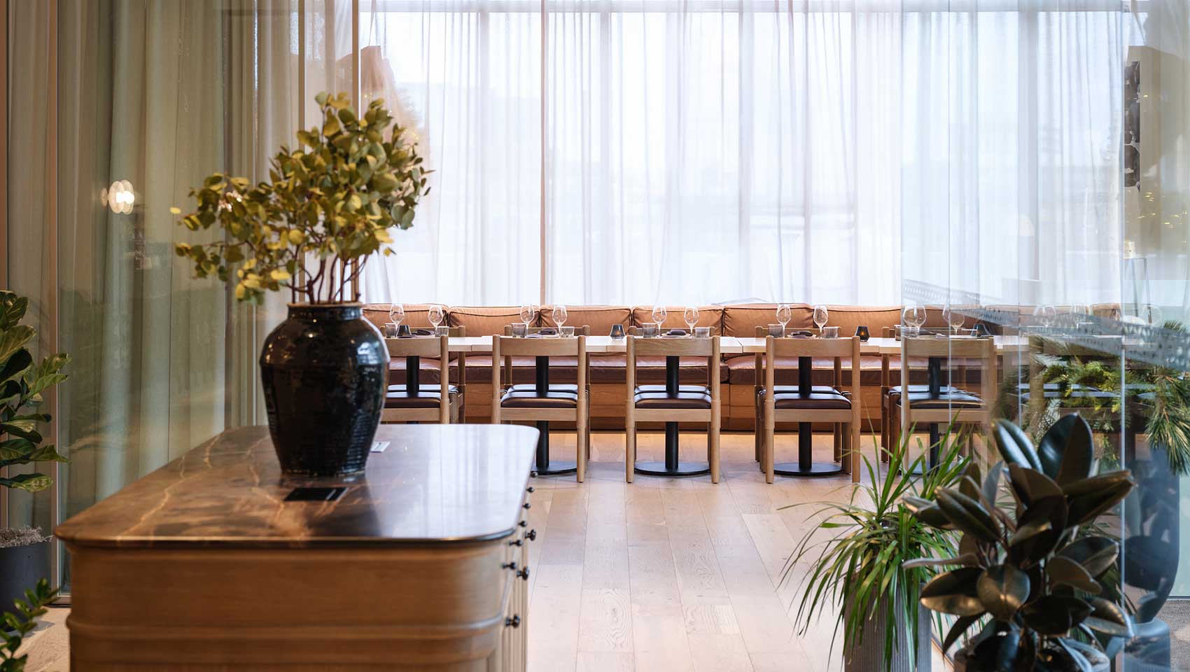 Looking from the lobby bar area into the dining room at table and chairs with wall of windows and soft curtains